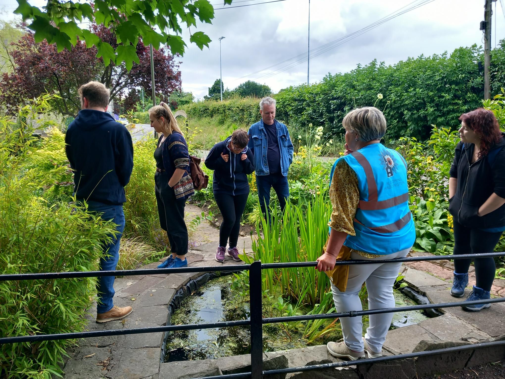 Six people gather around a small pond in a green, lush garden