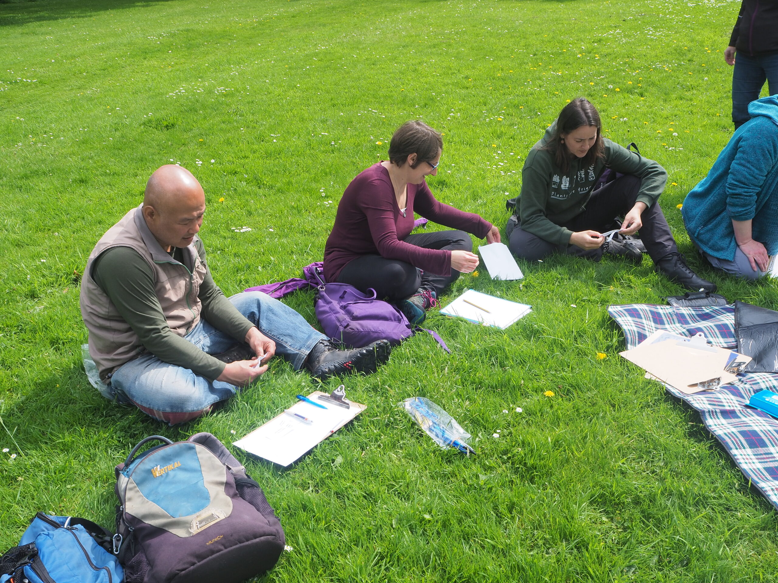 Three people sit on the grass with paper on the ground in front of them