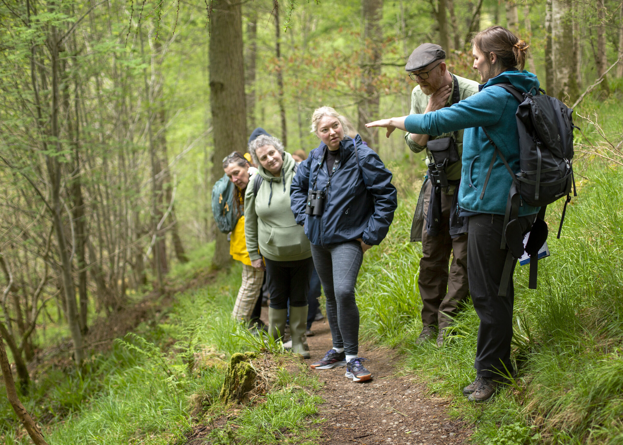 A group of people outside in a dense green forest, standing on the side of a path as one person points to the left off-camera